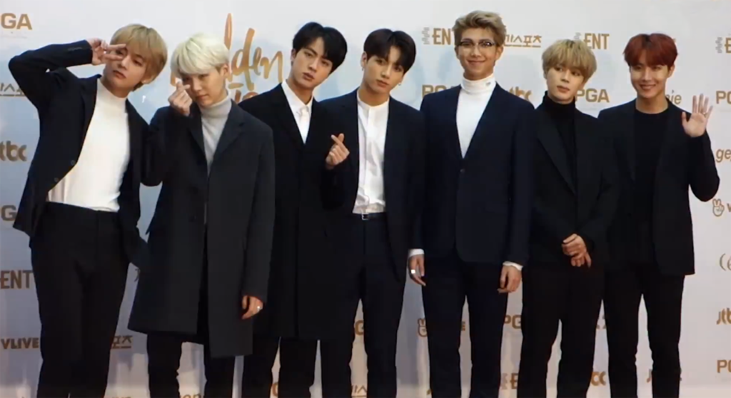 The seven members of the K-pop group BTS stand side-by-side on a red carpet, wearing dark suits and white tops, posing for the camera.