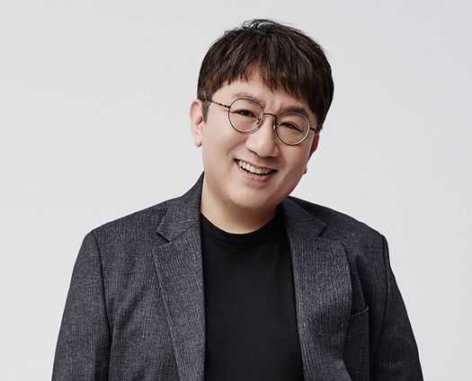 A headshot portrait of HYBE founder Bang Si-hyuk, smiling and wearing round glasses and a dark blazer over a black t-shirt against a plain white background.