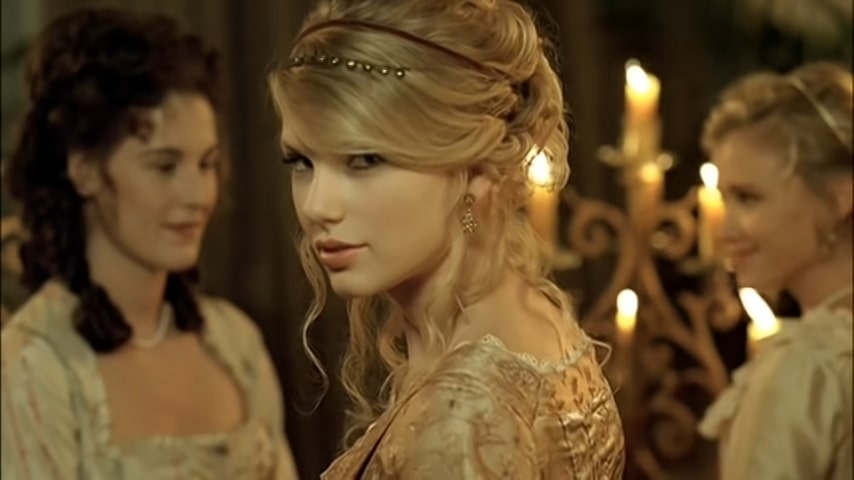 A cinematic still featuring a woman with blonde hair styled in an intricate updo with a delicate beaded headband, looking over her shoulder with a subtle smile. She is wearing a vintage-style gold brocade dress and elegant earrings. In the background, two other women in period clothing are partially visible, along with a warm-toned environment illuminated by several glowing candles on a decorative candelabra.