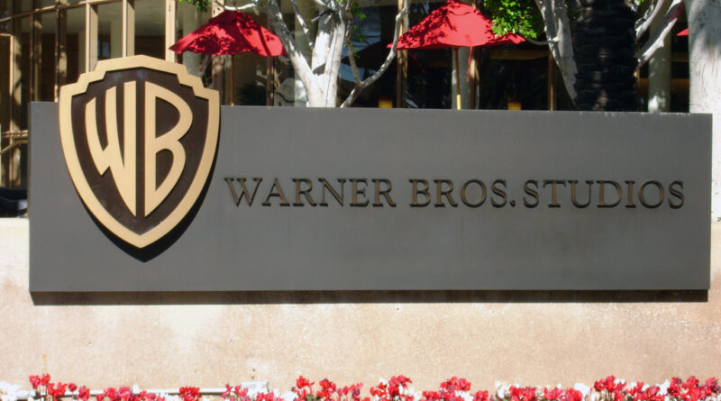 The entrance sign of Warner Bros. Studios featuring the golden WB shield logo, with red umbrellas and green trees in the background.