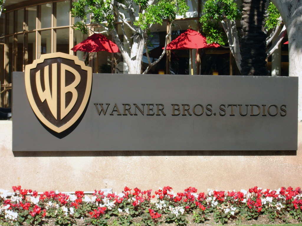 A medium shot of a modern dark gray sign bearing the words 'WARNER BROS. STUDIOS' in metallic lettering, accompanied by the classic golden 'WB' shield logo. The sign is placed outdoors, with a bed of red and white flowers at its base, and a glass-paneled building with green trees and red umbrellas visible behind it.