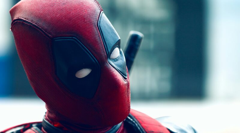 A close-up side profile of the character Deadpool in his signature red and black tactical suit, looking toward the right with a blurred background.