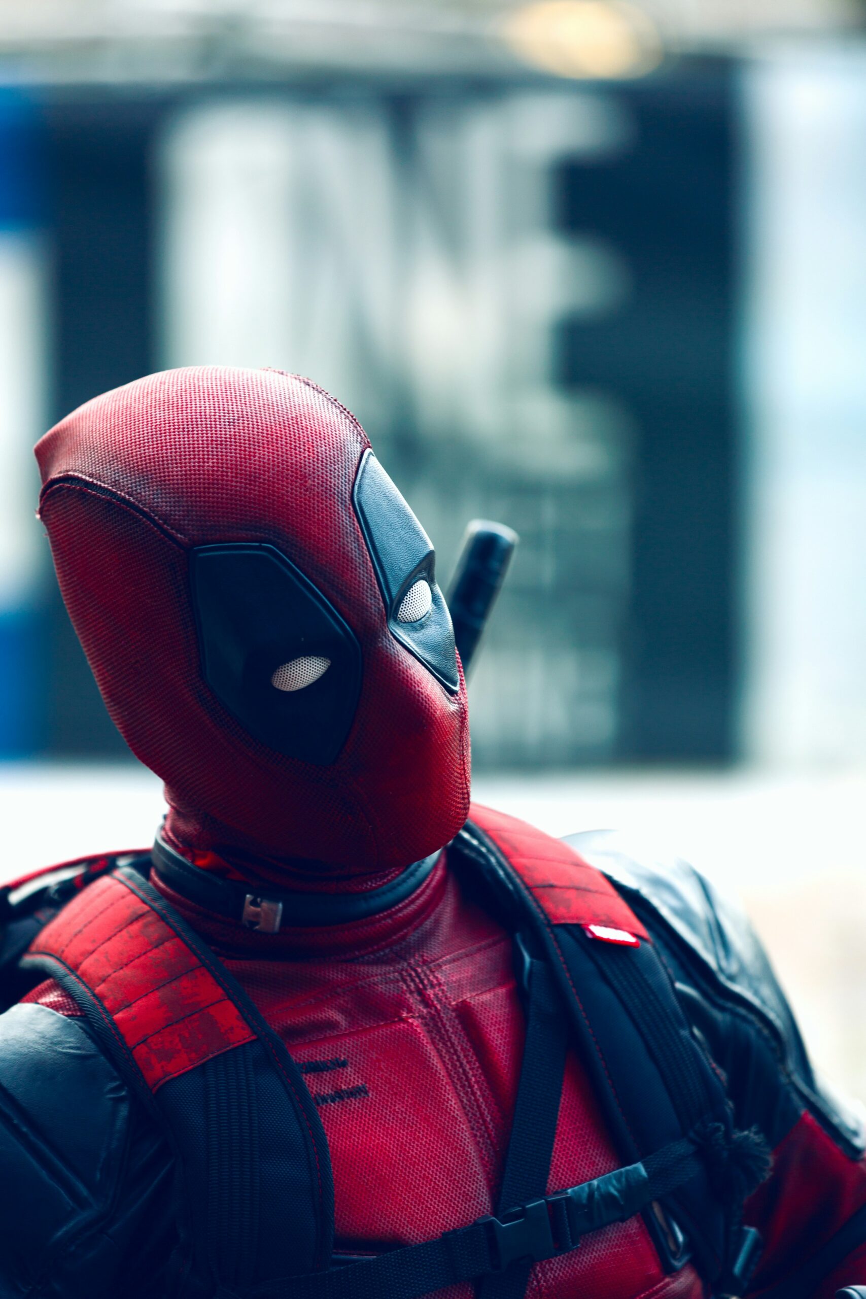 A close-up side profile of the character Deadpool in his signature red and black tactical suit, looking toward the right with a blurred background.