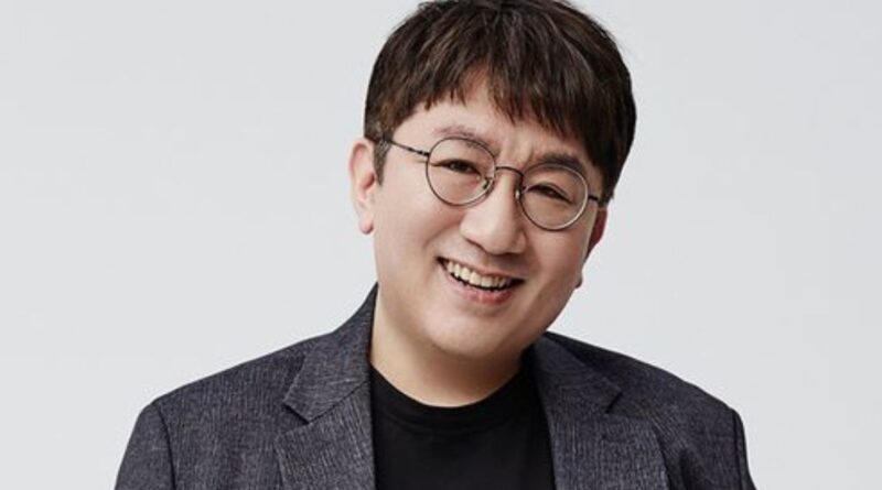 A headshot portrait of HYBE founder Bang Si-hyuk, smiling and wearing round glasses and a dark blazer over a black t-shirt against a plain white background.