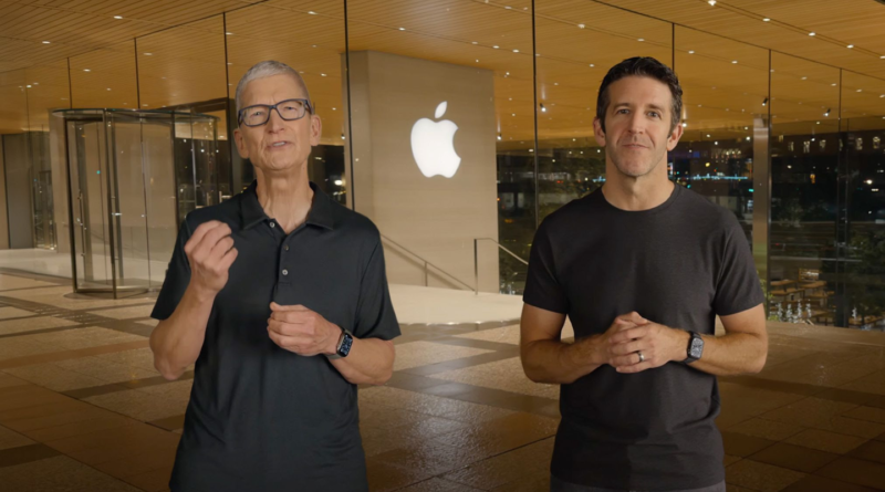 Tim Cook and John Ternus stand together outdoors in front of an Apple building with the company's illuminated logo in the background.