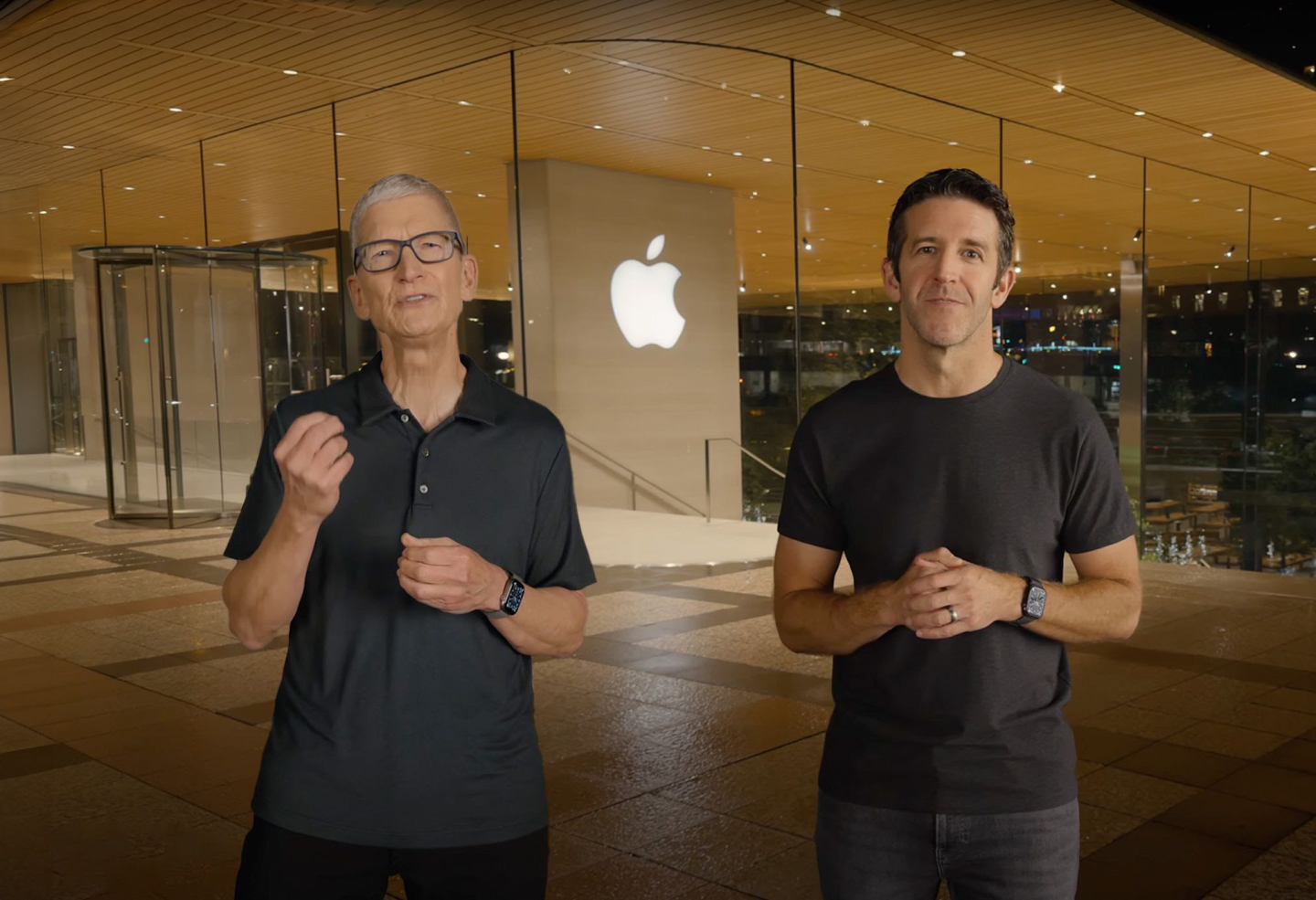 Tim Cook and John Ternus stand together outdoors in front of an Apple building with the company's illuminated logo in the background.
