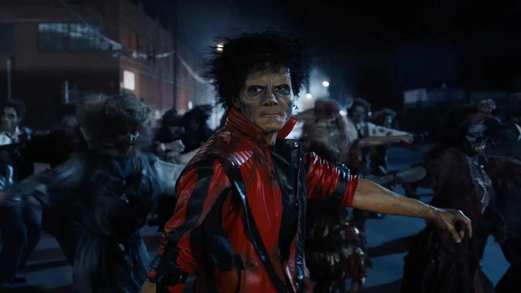A still photograph taken under dim blue lighting shows a group of dancers in ragged, 1980s-style clothing, including a man in a red and black leather jacket in the foreground, with pale makeup and wild, frizzy dark hair, standing in front of other performers with his arms outstretched to the sides. They are on an asphalt street with blurred industrial buildings and overhead lights in the background.