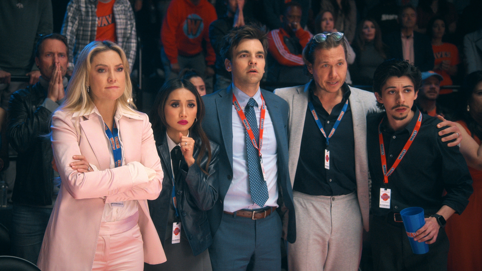 A group of five people, including Kate Hudson wearing a pink suit and other individuals with event lanyards, looking concerned and focused during an event.