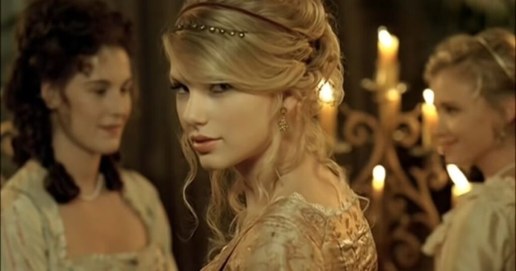  A cinematic still featuring a woman with blonde hair styled in an intricate updo with a delicate beaded headband, looking over her shoulder with a subtle smile. She is wearing a vintage-style gold brocade dress and elegant earrings. In the background, two other women in period clothing are partially visible, along with a warm-toned environment illuminated by several glowing candles on a decorative candelabra.