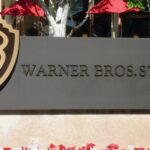 Warner Paramount Merger 2026 The entrance sign of Warner Bros. Studios featuring the golden WB shield logo, with red umbrellas and green trees in the background.