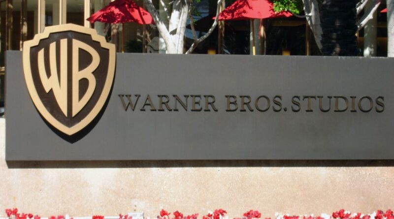 The entrance sign of Warner Bros. Studios featuring the golden WB shield logo, with red umbrellas and green trees in the background.