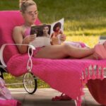 A young woman with blonde hair, wearing pink sunglasses and a pink outfit, lounging on a pink towel-covered chair and reading a magazine outdoors.