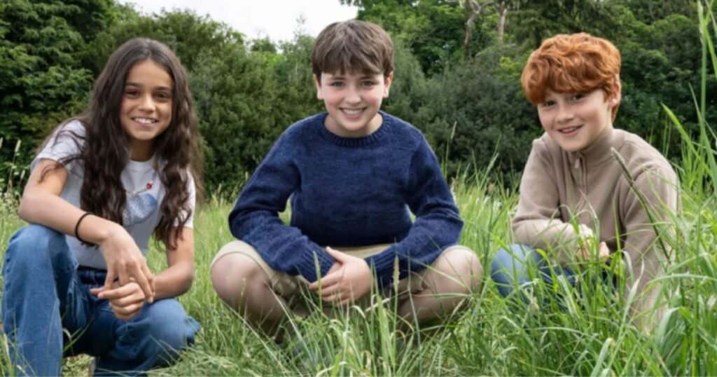 A color photograph of three young people sitting closely together outdoors in tall green grass, posing for the camera with smiles. The person on the left is a girl with long dark hair, wearing a white graphic t-shirt and blue jeans, looking down. The person in the center is a boy with brown hair in a navy blue knitted sweater and khaki shorts. The person on the right is a boy with curly reddish hair, wearing a light tan zipped sweater. Behind them is a large hedge of green bushes. The lighting is natural daylight.