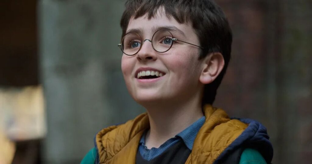 A close-up portrait of a young boy with dark hair and round glasses, smiling upward with an excited expression, wearing a yellow hooded vest over a blue shirt.