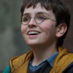 A close-up portrait of a young boy with dark hair and round glasses, smiling upward with an excited expression, wearing a yellow hooded vest over a blue shirt.