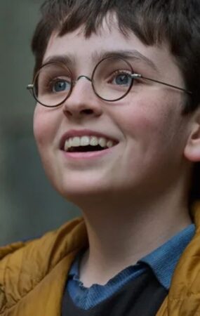 A close-up portrait of a young boy with dark hair and round glasses, smiling upward with an excited expression, wearing a yellow hooded vest over a blue shirt.
