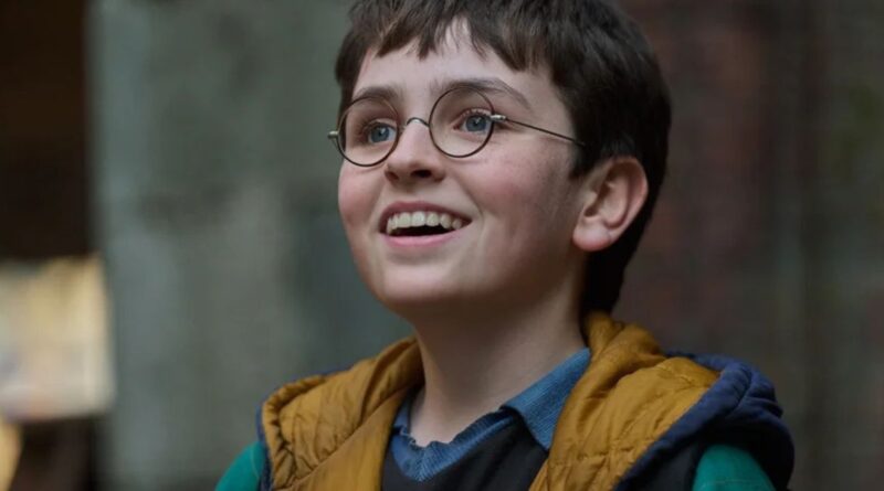 A close-up portrait of a young boy with dark hair and round glasses, smiling upward with an excited expression, wearing a yellow hooded vest over a blue shirt.