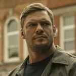 Close-up of Alan Ritchson as Jack Reacher, looking off-camera with a serious expression against a blurred urban building background.
