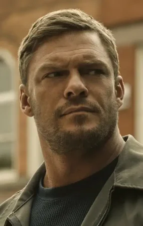 Close-up of Alan Ritchson as Jack Reacher, looking off-camera with a serious expression against a blurred urban building background.