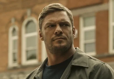 Close-up of Alan Ritchson as Jack Reacher, looking off-camera with a serious expression against a blurred urban building background.