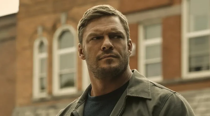 Close-up of Alan Ritchson as Jack Reacher, looking off-camera with a serious expression against a blurred urban building background.