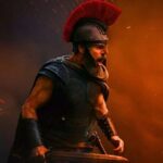 A powerful cinematic shot of a Greek warrior, portrayed by Matt Damon, wearing a black breastplate and a Spartan-style helmet with a large red crest. He is shouting in the heat of battle, holding a sword, amidst a backdrop of orange smoke, embers, and dark shadows.