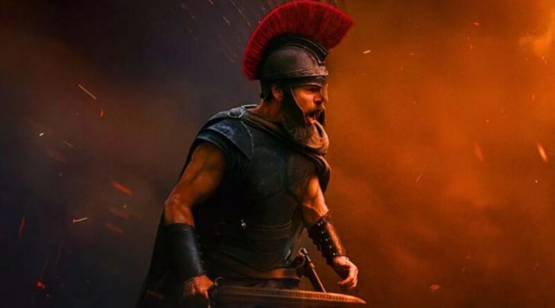 A powerful cinematic shot of a Greek warrior, portrayed by Matt Damon, wearing a black breastplate and a Spartan-style helmet with a large red crest. He is shouting in the heat of battle, holding a sword, amidst a backdrop of orange smoke, embers, and dark shadows.