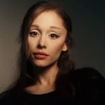 Close-up portrait of singer Ariana Grande looking at the camera with dark hair and a neutral expression.