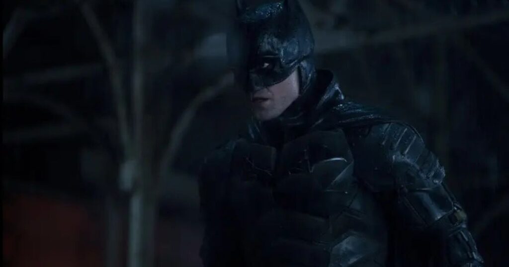A medium shot of Robert Pattinson as Batman, wearing his battle-worn tactical suit and cowl in a dark, rainy environment. He is looking off to the side with an intense and vigilant expression, his armor glistening from the rain.