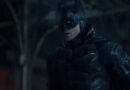 A medium shot of Robert Pattinson as Batman, wearing his battle-worn tactical suit and cowl in a dark, rainy environment. He is looking off to the side with an intense and vigilant expression, his armor glistening from the rain.