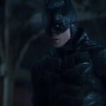 A medium shot of Robert Pattinson as Batman, wearing his battle-worn tactical suit and cowl in a dark, rainy environment. He is looking off to the side with an intense and vigilant expression, his armor glistening from the rain.
