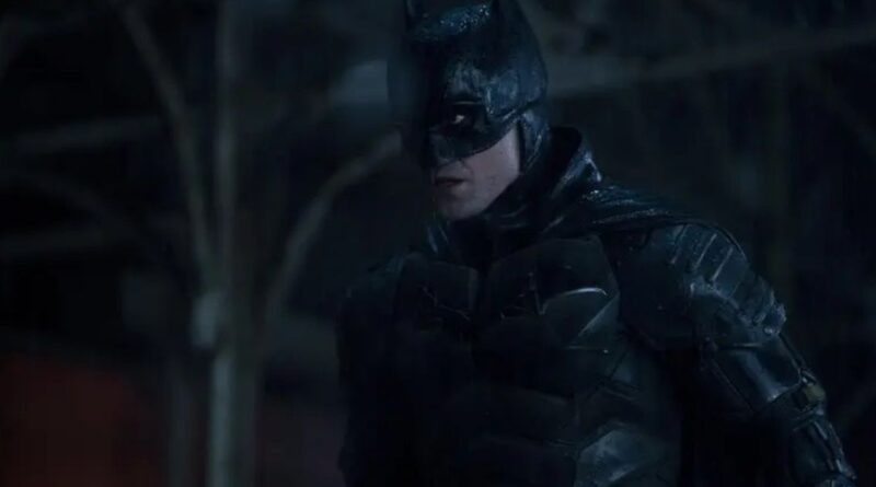 A medium shot of Robert Pattinson as Batman, wearing his battle-worn tactical suit and cowl in a dark, rainy environment. He is looking off to the side with an intense and vigilant expression, his armor glistening from the rain.