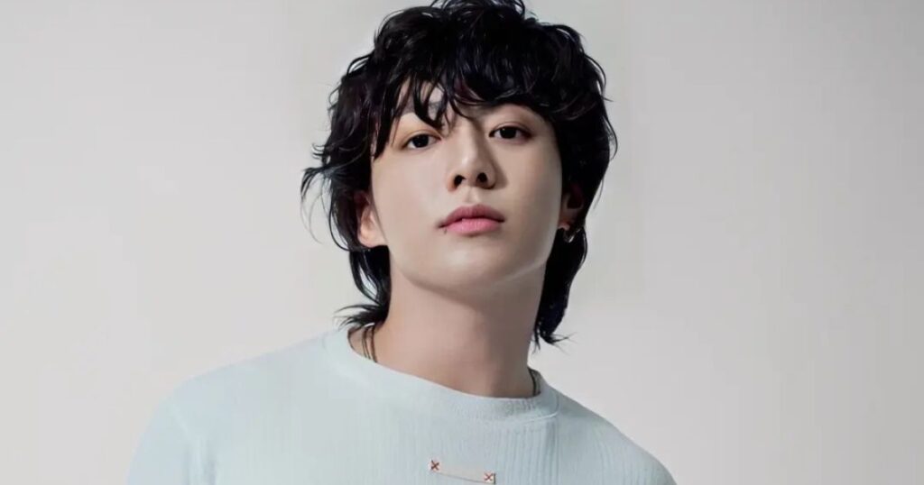 A clean, high-resolution studio portrait of South Korean singer Jungkook. He is looking directly at the camera with a calm expression, featuring his signature wavy black hair and a small lip piercing. He is wearing a simple, light-colored ribbed sweater against a neutral, off-white background.