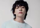 A clean, high-resolution studio portrait of South Korean singer Jungkook. He is looking directly at the camera with a calm expression, featuring his signature wavy black hair and a small lip piercing. He is wearing a simple, light-colored ribbed sweater against a neutral, off-white background.
