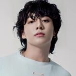 A clean, high-resolution studio portrait of South Korean singer Jungkook. He is looking directly at the camera with a calm expression, featuring his signature wavy black hair and a small lip piercing. He is wearing a simple, light-colored ribbed sweater against a neutral, off-white background.