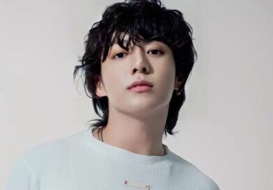 A clean, high-resolution studio portrait of South Korean singer Jungkook. He is looking directly at the camera with a calm expression, featuring his signature wavy black hair and a small lip piercing. He is wearing a simple, light-colored ribbed sweater against a neutral, off-white background.