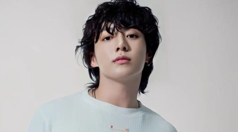 A clean, high-resolution studio portrait of South Korean singer Jungkook. He is looking directly at the camera with a calm expression, featuring his signature wavy black hair and a small lip piercing. He is wearing a simple, light-colored ribbed sweater against a neutral, off-white background.