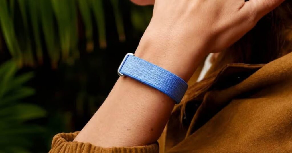 A close-up shot of a person's wrist wearing a light blue woven fabric smart band with a silver buckle. The person is wearing a brown jacket, and the background shows blurred green foliage, focusing on the texture and fit of the wearable device.