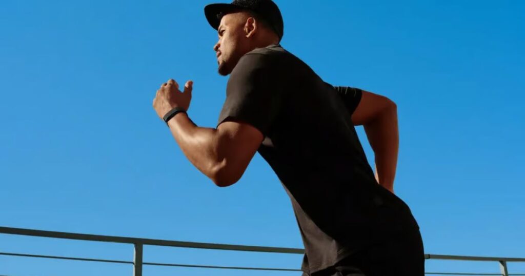 A side-view photograph of a man with a beard and a black cap running outdoors under a clear blue sky. He is wearing a black t-shirt and a sleek black smart band on his right wrist. The low-angle shot shows him in motion against a minimalist background with a simple railing.