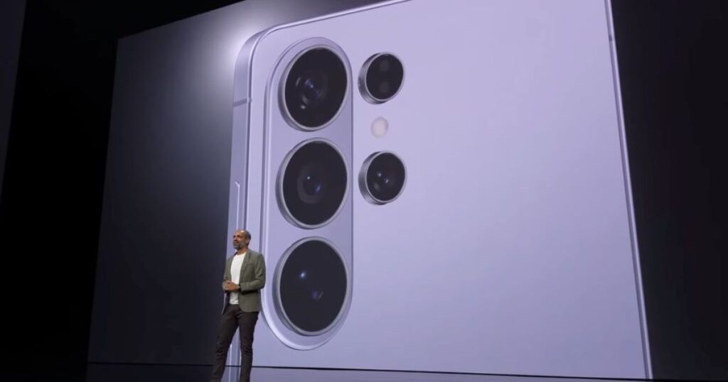 A wide shot of a tech keynote presentation stage where a presenter stands in front of a massive digital screen. The screen displays a close-up, high-detail view of a light purple Samsung Galaxy smartphone's rear camera island, featuring four distinct circular camera lenses and a flash.