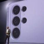 A wide shot of a tech keynote presentation stage where a presenter stands in front of a massive digital screen. The screen displays a close-up, high-detail view of a light purple Samsung Galaxy smartphone's rear camera island, featuring four distinct circular camera lenses and a flash.