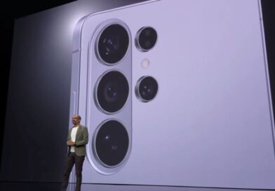 A wide shot of a tech keynote presentation stage where a presenter stands in front of a massive digital screen. The screen displays a close-up, high-detail view of a light purple Samsung Galaxy smartphone's rear camera island, featuring four distinct circular camera lenses and a flash.