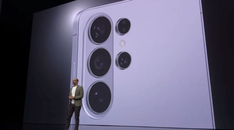 A wide shot of a tech keynote presentation stage where a presenter stands in front of a massive digital screen. The screen displays a close-up, high-detail view of a light purple Samsung Galaxy smartphone's rear camera island, featuring four distinct circular camera lenses and a flash.