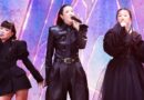 Three female singers in black outfits performing on a brightly lit stage, each holding a microphone.