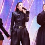 Three female singers in black outfits performing on a brightly lit stage, each holding a microphone.