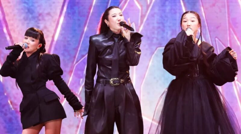 Three female singers in black outfits performing on a brightly lit stage, each holding a microphone.