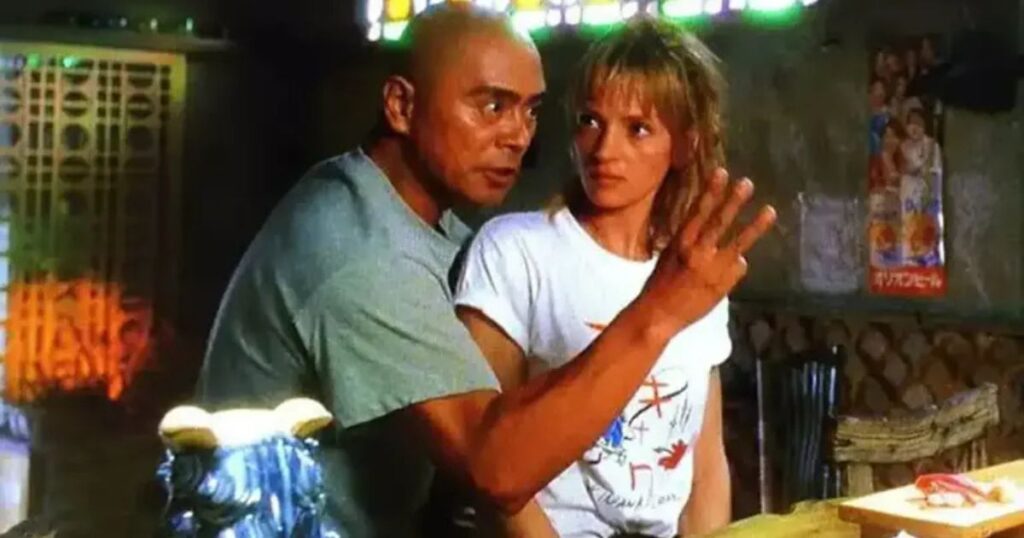A candid production still from the movie Kill Bill: Volume 1 featuring Japanese actor Kenji Ohba and actress Uma Thurman. Ohba, with a shaved head and wearing a grey t-shirt, is pointing off-camera while Thurman, in a white t-shirt, looks on with a curious expression. The background shows the rustic interior of a Japanese izakaya.
