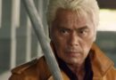 A close-up, dramatic shot of veteran actor Kenji Ohba with blonde hair, wearing a brown leather jacket. He has an intense, focused expression as a silver metallic blade is partially visible in the foreground.