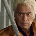 A close-up, dramatic shot of veteran actor Kenji Ohba with blonde hair, wearing a brown leather jacket. He has an intense, focused expression as a silver metallic blade is partially visible in the foreground.