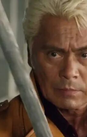 A close-up, dramatic shot of veteran actor Kenji Ohba with blonde hair, wearing a brown leather jacket. He has an intense, focused expression as a silver metallic blade is partially visible in the foreground.